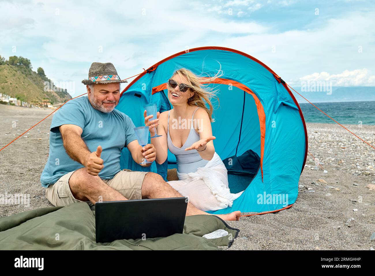 Coppia che si diverte, beve vino e parla con gli amici in videochiamata nel laptop in un picnic vicino a una tenda sul mare. Vacanze estive sulla spiaggia di Outd Foto Stock