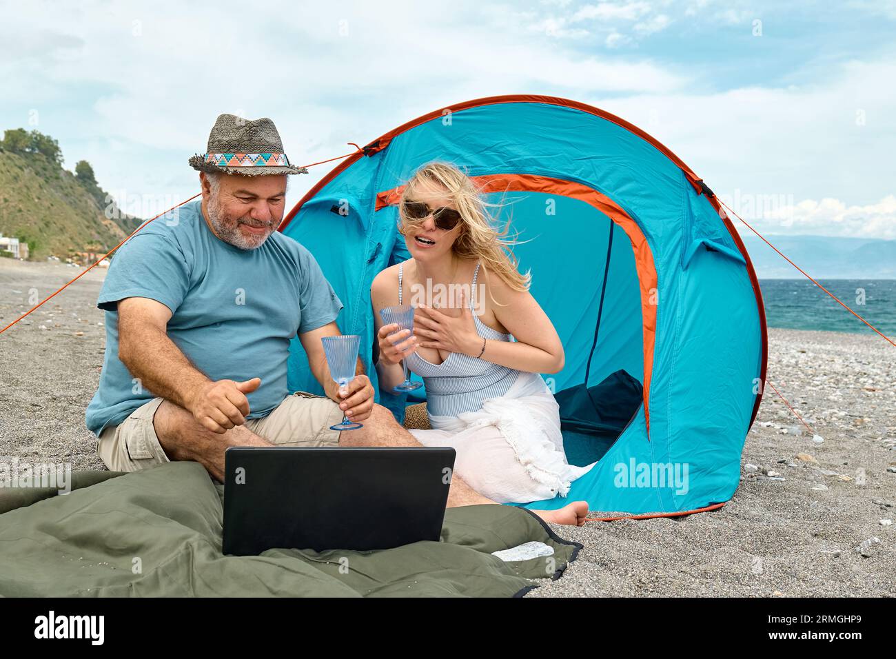 Coppia che si diverte, beve vino e parla con gli amici in videochiamata nel laptop in un picnic vicino a una tenda sul mare. Vacanze estive sulla spiaggia di Outd Foto Stock