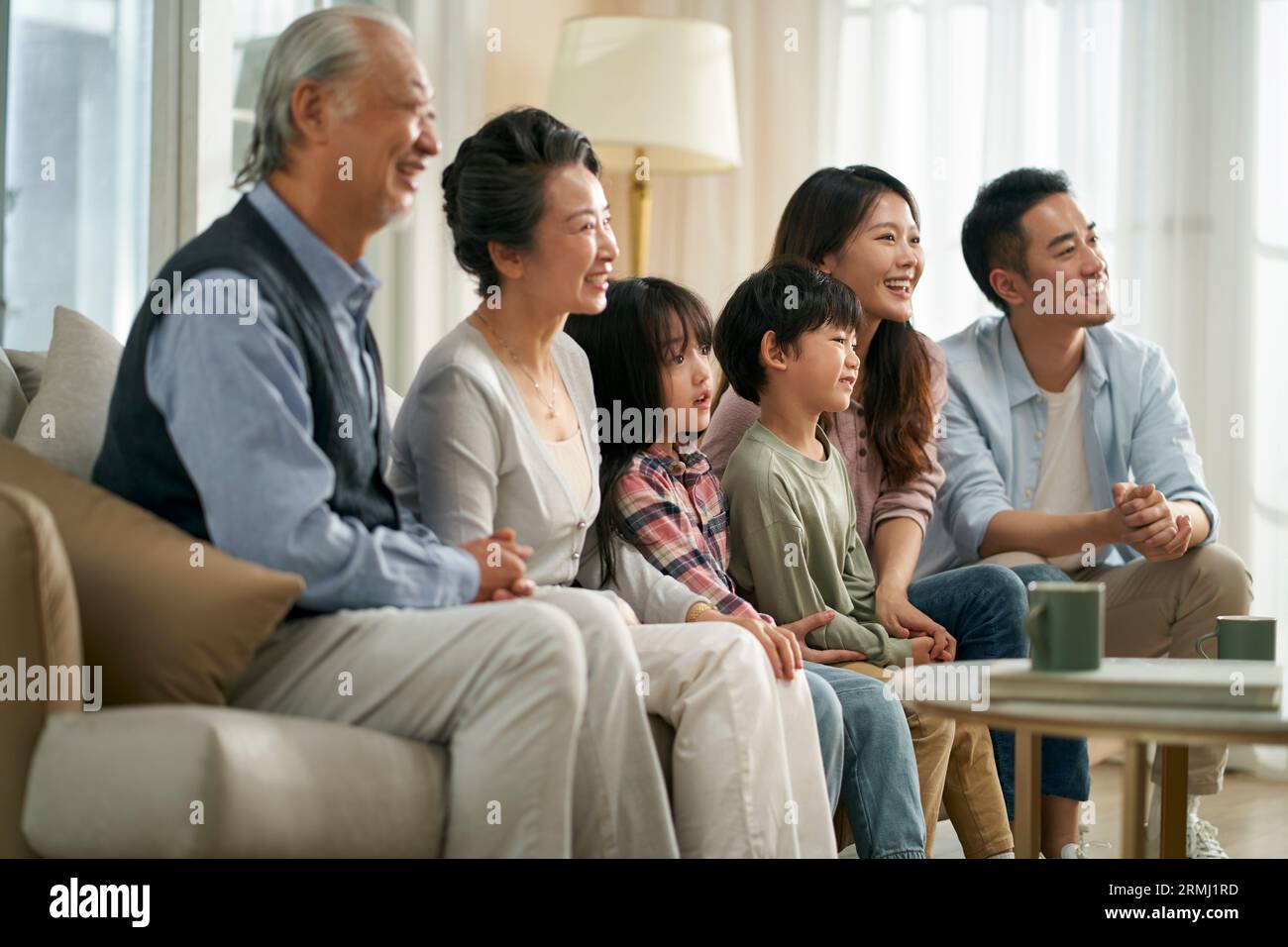 famiglia asiatica di tre generazioni seduta sul divano a casa a guardare la tv insieme felice e sorridente Foto Stock