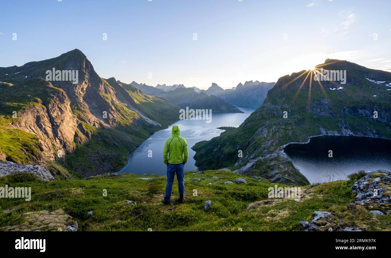Alpinista che guarda al paesaggio montano con le vette appuntite e il fiordo Forsfjorden con il villaggio di Vindstad, stella del sole all'alba Foto Stock