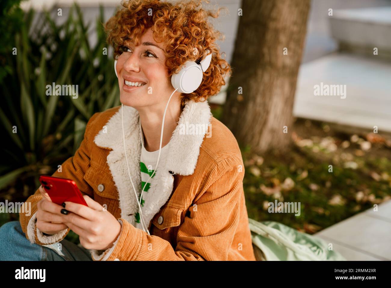 Donna di colore bianco medio con capelli ricci che usa il cellulare e le cuffie mentre cammina per la città Foto Stock