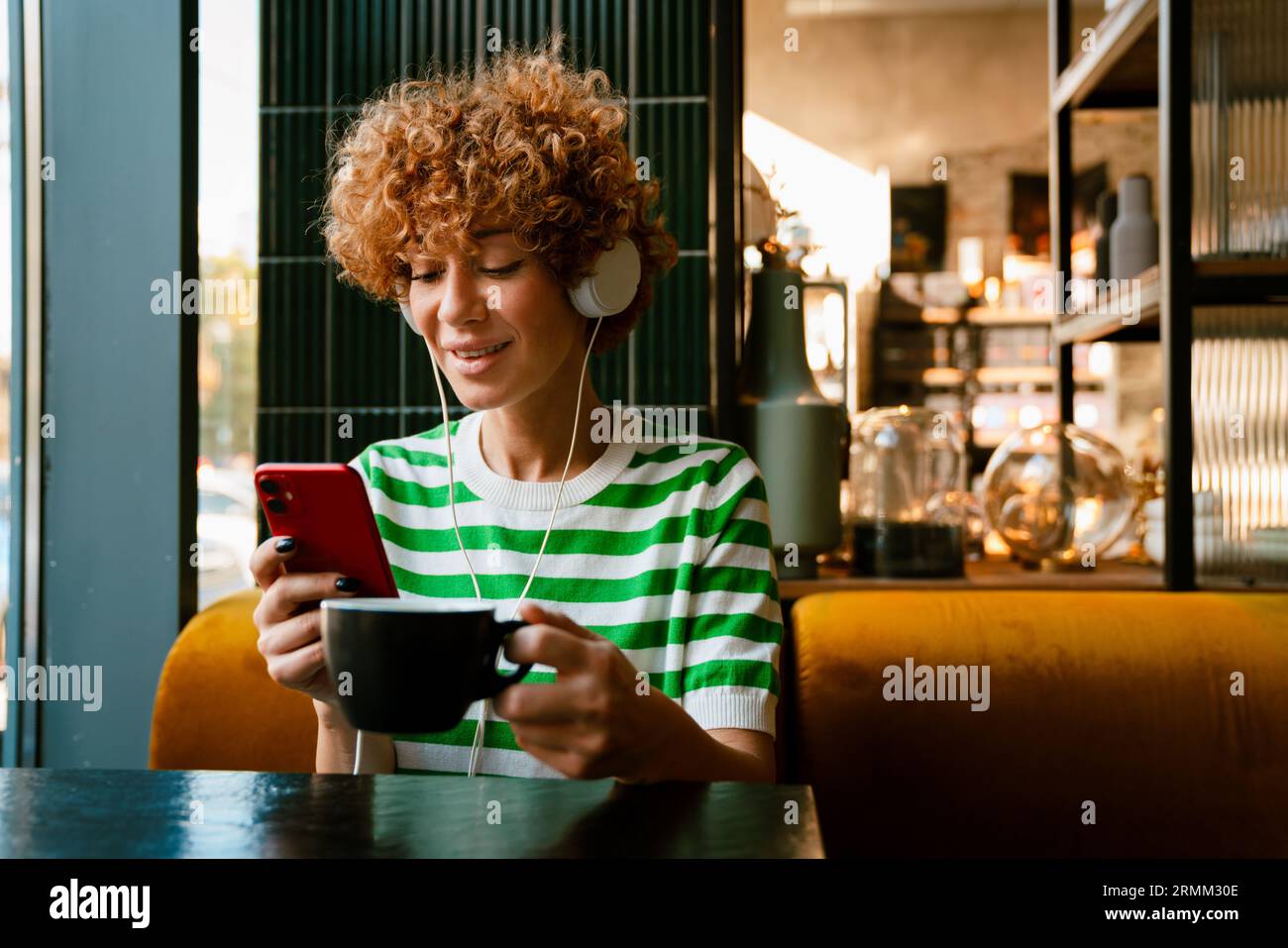 Donna sorridente di mezza età con le cuffie che usa il telefono cellulare mentre beve un caffè in una caffetteria al chiuso Foto Stock