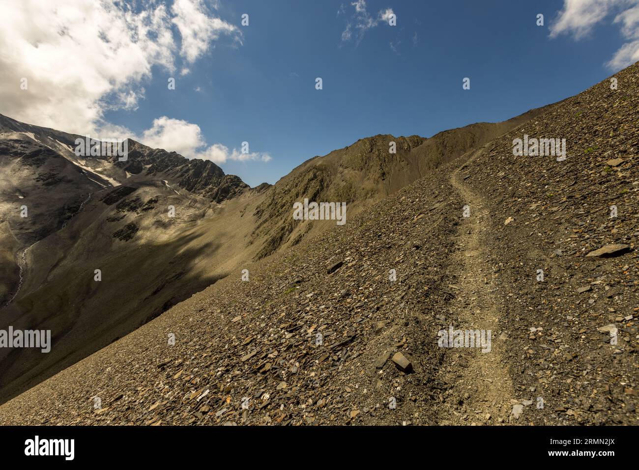 Un sentiero stretto attraversa una ripida pendenza di detriti di ardesia scura durante l'ascesa al Passo Atsunta nella municipalità di Akhmeta, regione di Kakheti, Georgia. Cresta rocciose con macchie di neve si ergono a sinistra. Sotto di esse, una pendenza scende in una valle ombrosa. Nuvole bianche si muovono nel cielo blu. L'ascesa si snoda a zigzag su detriti. Il Passo Atsunta collega le regioni di Tusheti e Khevsureti. È alto più di 3.400 metri. I semi-nomadi Tushetiani vivono nei villaggi di montagna solo da luglio a settembre. Passano il resto dell'anno nella valle più bassa di Alvani. Foto Stock