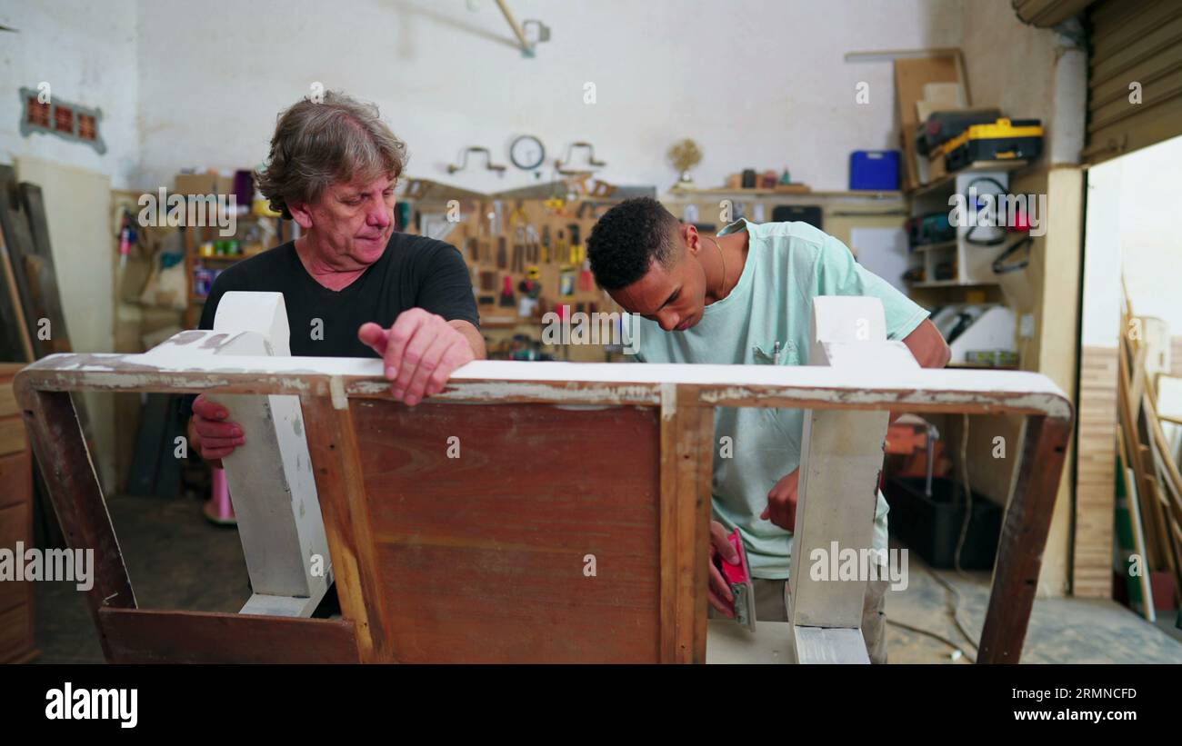 Giovani e maestri falegnami impegnati con il lavoro all'interno di un'officina di falegnameria. Falegname più anziano ed esperto che guida il dipendente alla lucidatura del legno Foto Stock