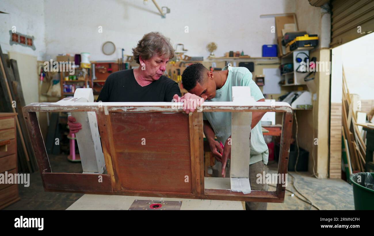Giovani e maestri falegnami impegnati con il lavoro all'interno di un'officina di falegnameria. Falegname più anziano ed esperto che guida il dipendente alla lucidatura del legno Foto Stock
