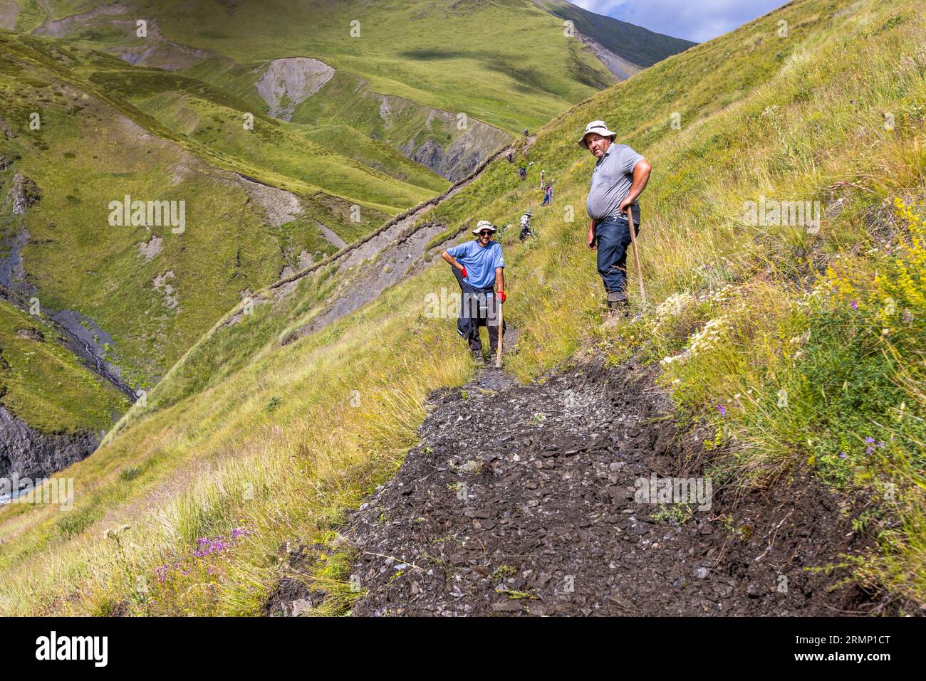 Due operai con attrezzi a manico lungo si trovano su una pista di terra appena rialzata su una ripida pendenza erbosa sulla via dalle praterie di Kvakhidi al passo Atsunta nella municipalità di Akhmeta, regione di Kakheti, Georgia. L'uomo a destra indossa una polo grigia, jeans e un cappello da sole chiaro. Si appoggia sul manico. L'uomo a sinistra indossa una maglietta blu, occhiali da sole, un cappello da sole e guanti da lavoro rossi. Ha una mano sul fianco. Più in alto sul pendio, uomini lavorano a intervalli lungo il sentiero. In basso a sinistra, un fiume scorre ai piedi di una gola. Fiori selvatici gialli, bianchi e viola sbocciano accanto Foto Stock