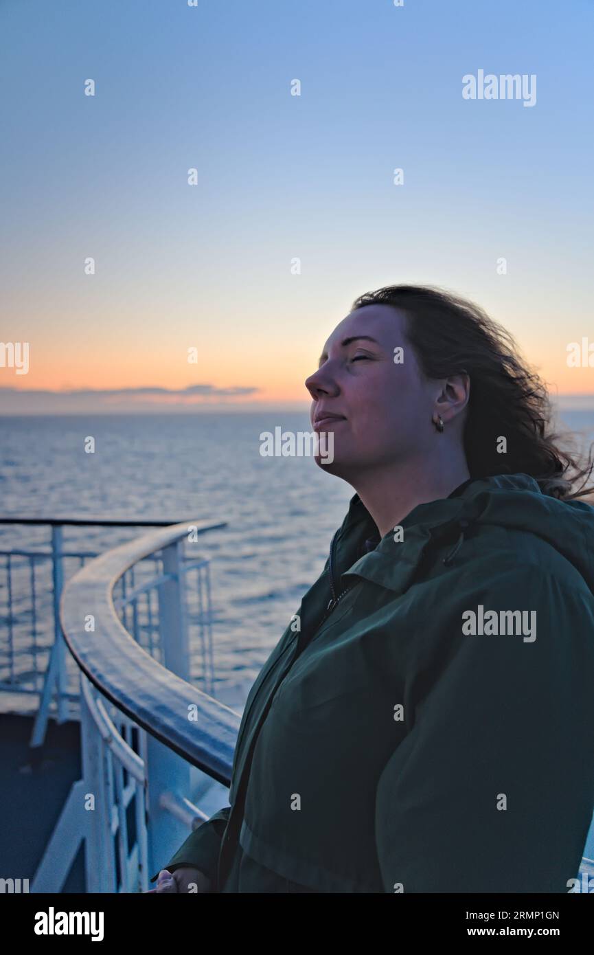 Bella donna che si rilassa sul balcone della barca con gli occhi chiusi e il vento tra i capelli. Tramonto brillante sulla costa scandinava del mare Foto Stock