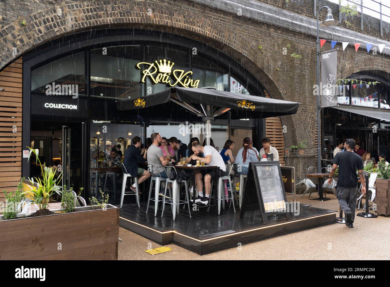Persone sedute e bevute fuori sotto la pioggia fuori dal Roti King situato sotto gli archi del Circus West Village su Arches Lane. Londra, Regno Unito Foto Stock