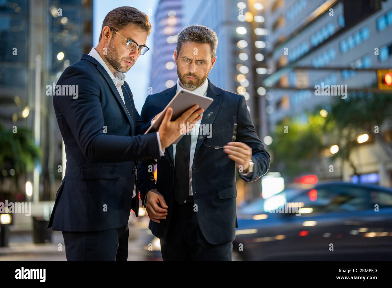 Gli uomini d'affari controllano le loro e-mail e esaminano documenti importanti su tablet sullo sfondo notturno della città. Due uomini d'affari usano i tablet per lavorare all'aperto Foto Stock