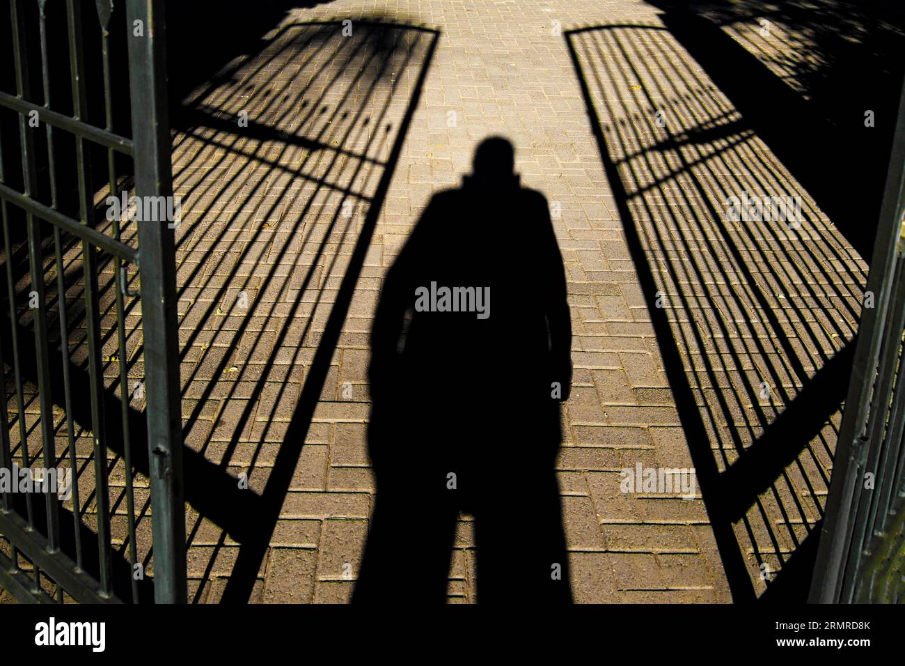 Ombra di una persona in piedi, gambe separate e mani in tasche, tra due cancelli in ferro battuto semi-aperti Foto Stock