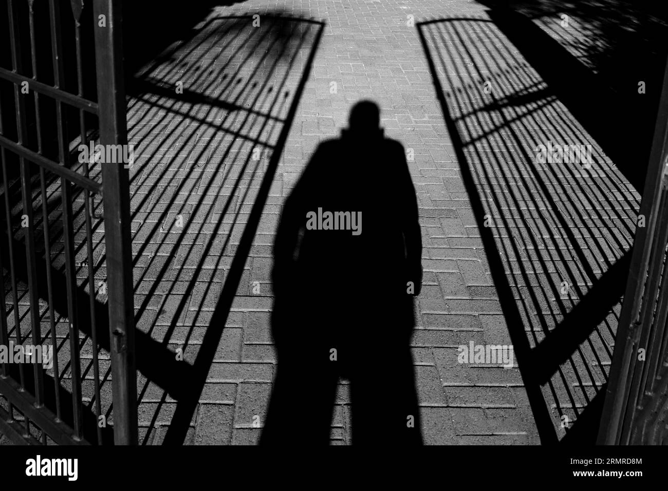 Ombra di una persona in piedi, gambe separate e mani in tasche, tra due cancelli in ferro battuto semi-aperti Foto Stock
