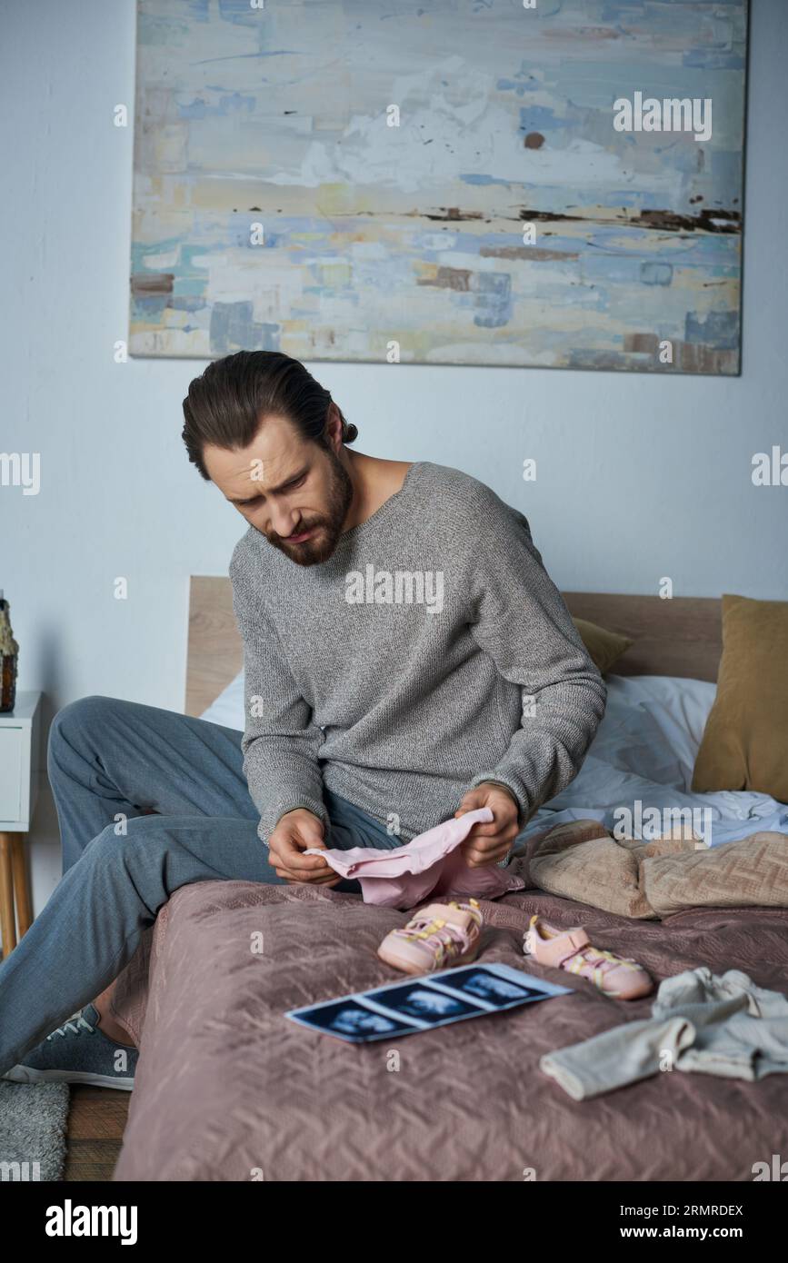 perdita, uomo depresso seduto sul letto vicino ai vestiti del bambino, ecografia, concetto di aborto spontaneo Foto Stock