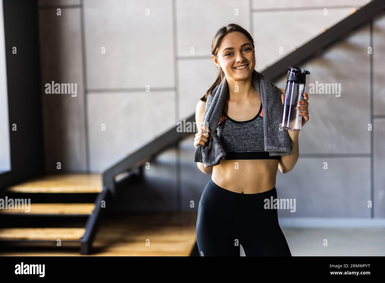 Ritratto di bella donna sorridente sportiva in abbigliamento sportivo e asciugamano bianco sul collo tenere shaker con bevanda sana, proteine del siero di latte, acqua fresca o caffè Foto Stock