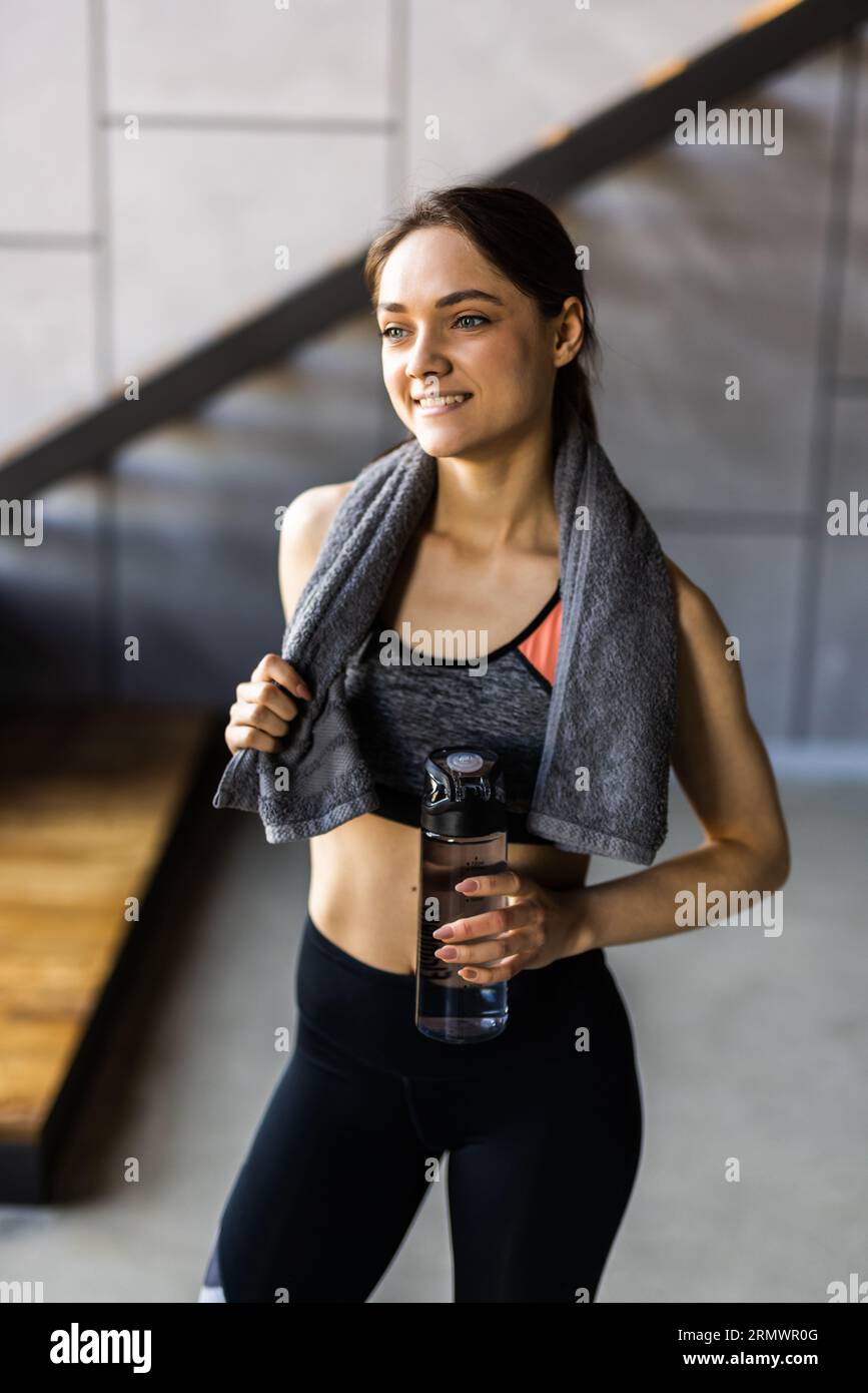 Ritratto di bella donna sorridente sportiva in abbigliamento sportivo e asciugamano bianco sul collo tenere shaker con bevanda sana, proteine del siero di latte, acqua fresca o caffè Foto Stock
