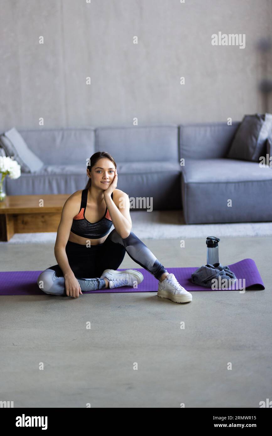 Soddisfatta blogger europea bella e giovane bionda che si riposa dopo l'allenamento a casa con l'acqua, donna prende una pausa sul tappeto nella palestra domestica. Vita sana Foto Stock
