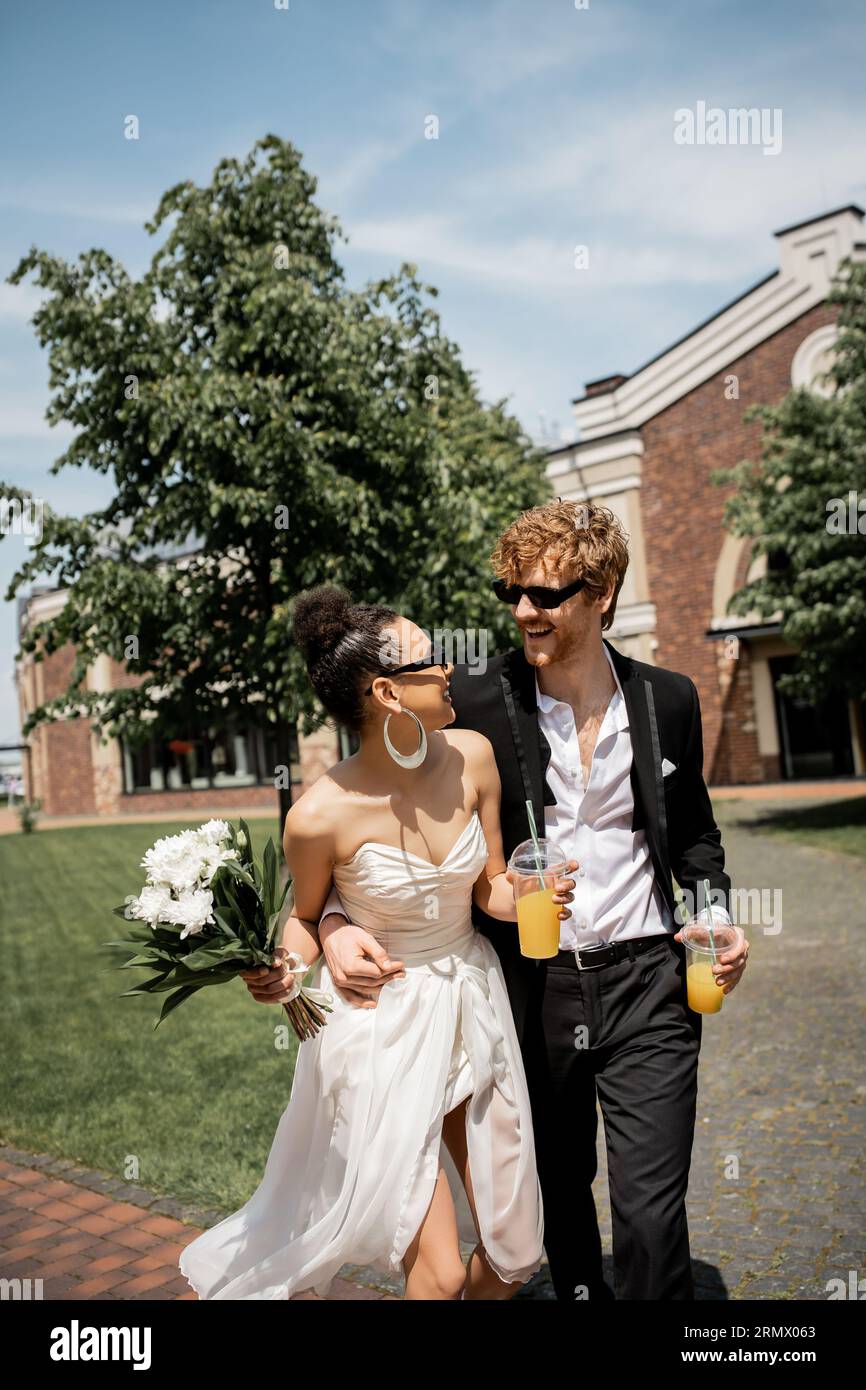 coppia variegata con succo d'arancia e fiori che sorridono l'uno all'altro, matrimonio per strada Foto Stock
