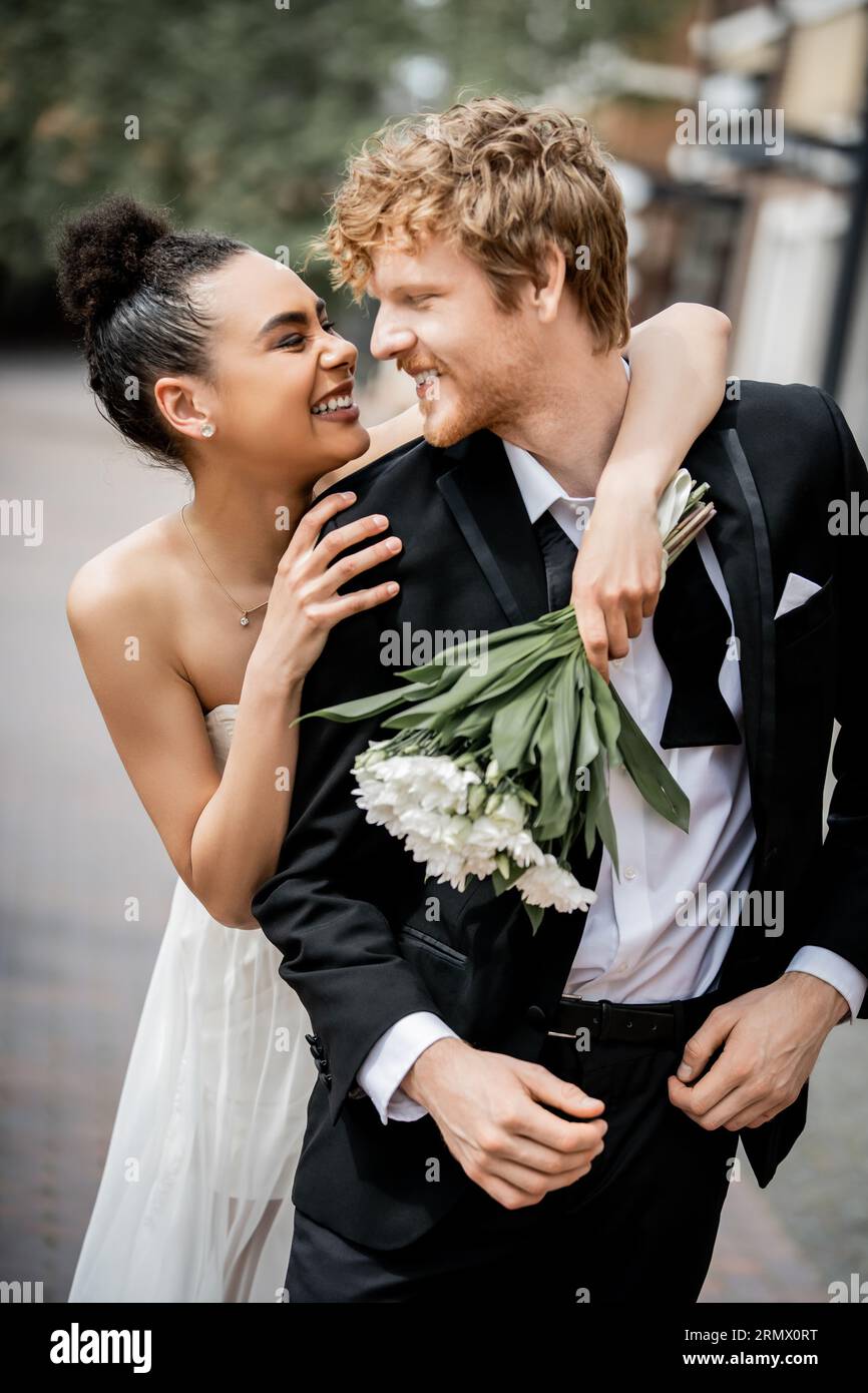 celebrazione di matrimoni all'aperto, eccitata sposa afroamericana con bouquet che abbraccia lo sposo di rosse Foto Stock
