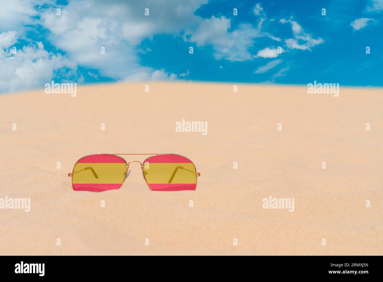 Gli occhiali da sole con occhiali in forma di bandiera della Spagna giacciono sulla sabbia contro il cielo blu. Concetto di vacanze estive, viaggi e turismo in Spagna. Riposo estivo Foto Stock