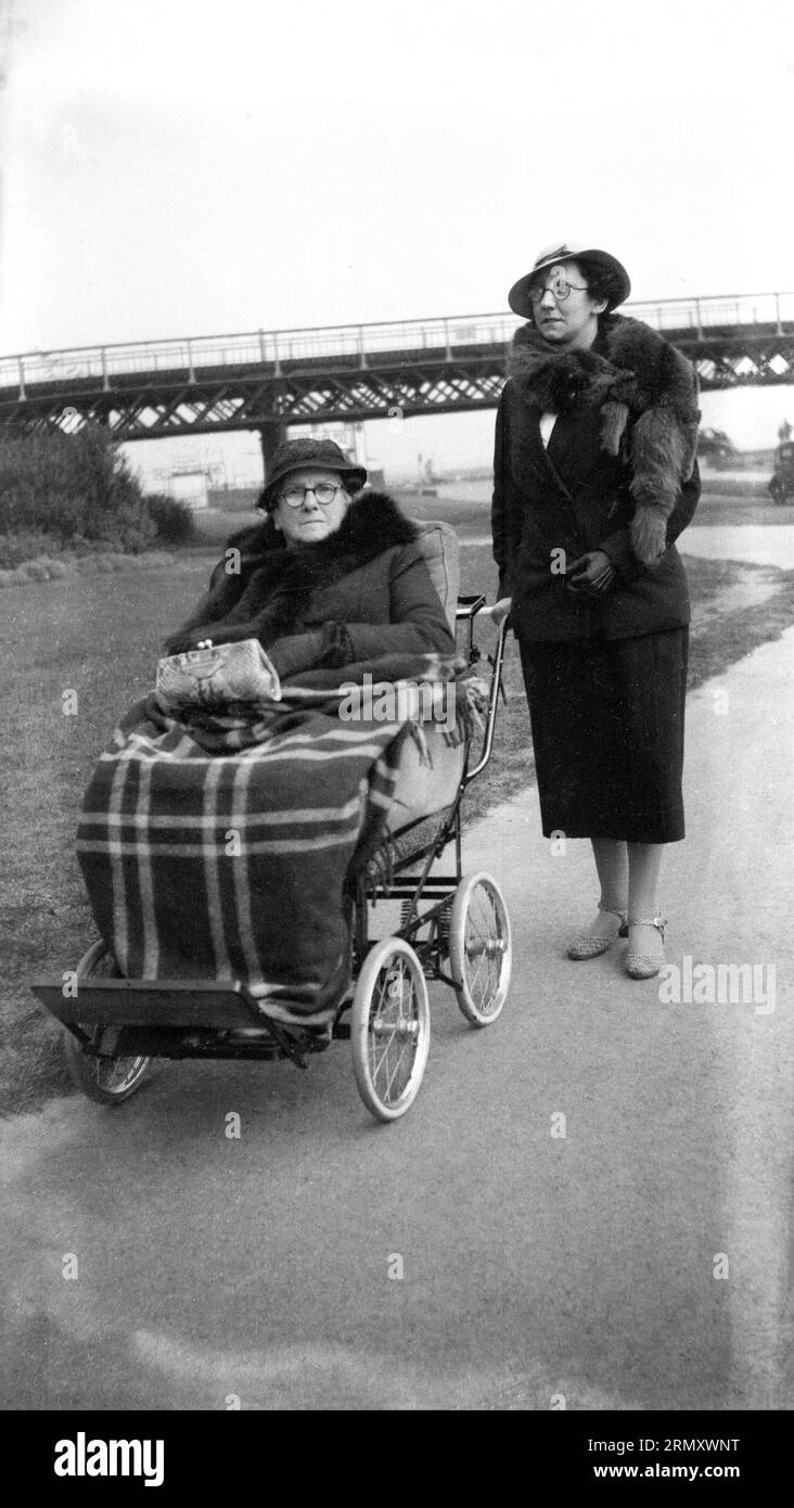 Anni '1930, storica, fuori su un sentiero, una signora che porta una signora anziana, seduto su un grande passeggino o un passeggino con una coperta sopra di lei, fuori per una passeggiata e un po' di aria fresca, Inghilterra, Regno Unito. I passeggini per adulti devono essere leggeri per facilitare il trave e allo stesso tempo abbastanza forti da poter accontentare il corpo adulto. Foto Stock