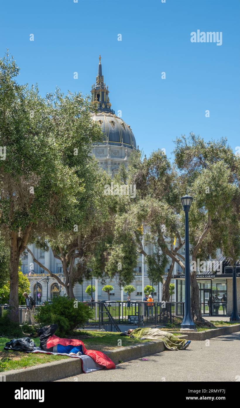 San Francisco, CA, USA - July 12, 2023: Homeless people sleep on Civic Center Plaza in front of City Hall. Its dome peeks over tree foliage under blue Foto Stock
