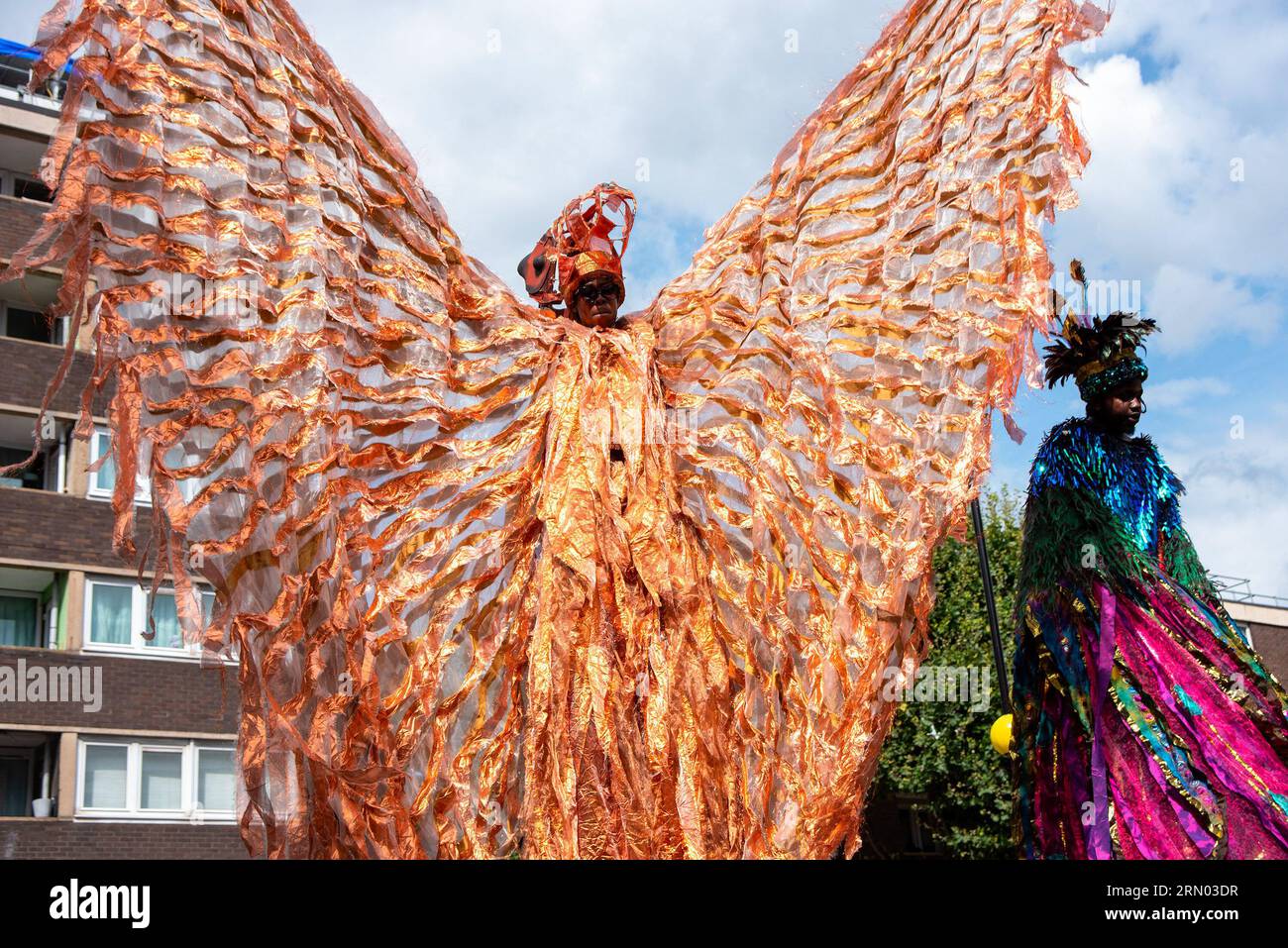 Un ballerino mette in onda il suo costume mentre cammina su palafitte durante la sfilata del Carnevale di Notting Hill a Londra. Il Carnevale di Notting Hill è uno dei più grandi festival di strada del mondo. È iniziato nel 1966, ma ha avuto origine con il Carnevale caraibico organizzato nel 1959 con la comunità di immigrati da Trinidad e Tobago. Quest'anno il Carnevale di Notting Hill segna il 50° anniversario dell'introduzione dei sistemi audio e delle band Mas. Inoltre segna il 75° anniversario dell'arrivo dei passeggeri dell'Empire Windrush nel Regno Unito. Foto Stock