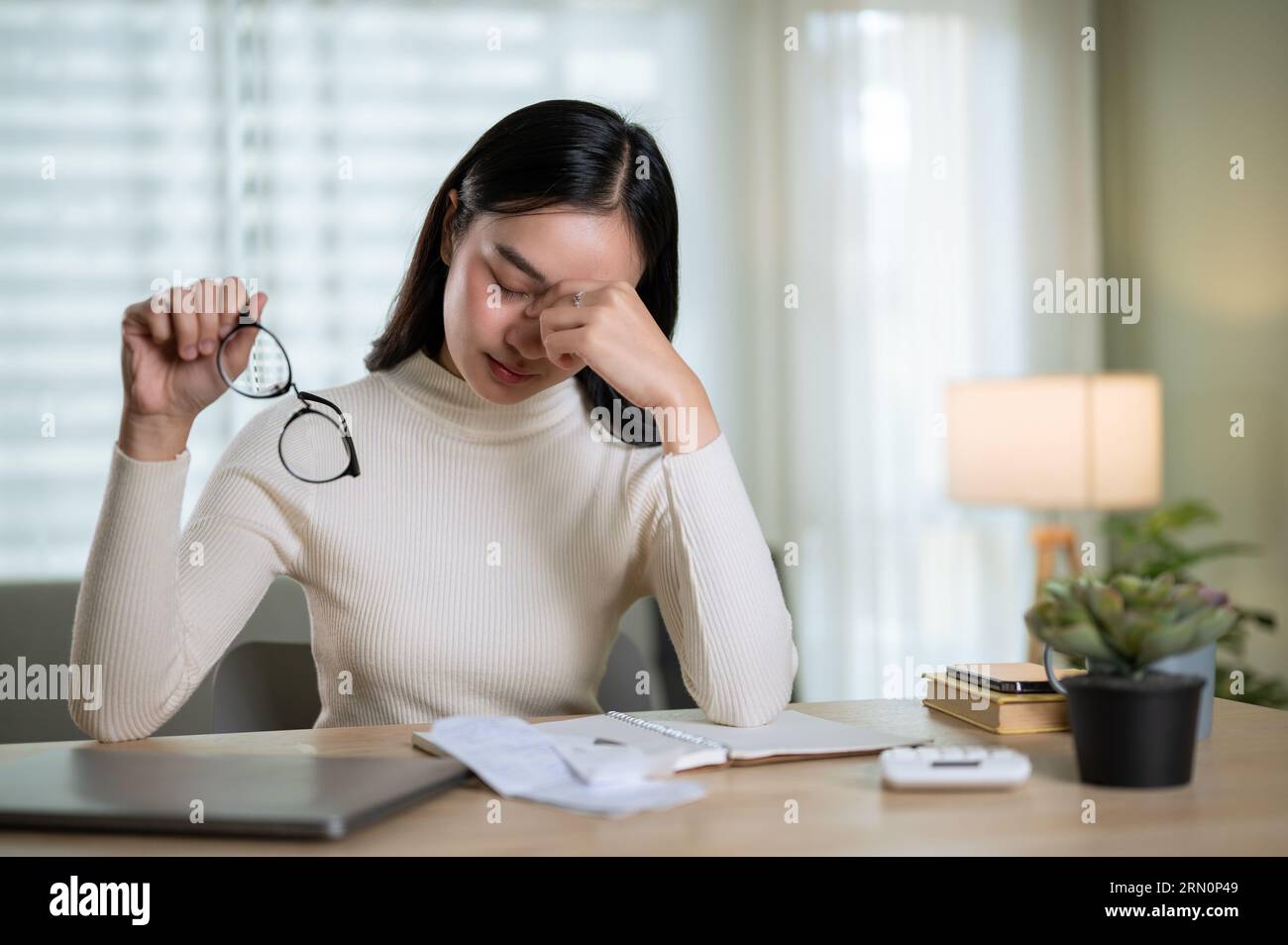 Una giovane donna asiatica, stressata, sta calcolando le spese mensili della sua famiglia, sentendosi frustrata, avendo mal di testa e preoccupata per i problemi finanziari Foto Stock