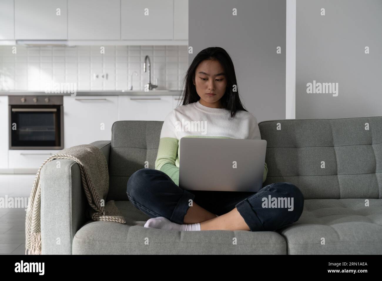 Una donna coreana professionista concentrata siede sul divano con un computer portatile e scrive testo in remoto Foto Stock