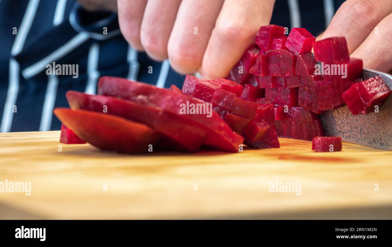 barbabietola fresca tagliata a cubetti con un coltello da cucina su un tagliere di legno con un grembiule sullo sfondo, vegetariano, vegano, preparazione del pasto Foto Stock