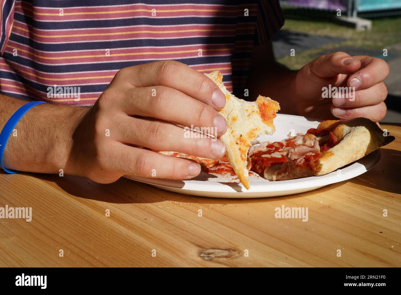 Ragazzo che mangia pizza con le mani nel ristorante all'aperto durante la giornata di sole. Ha una t-shirt rossa con cinturini e un bracciale in gomma blu sulla mano destra. Foto Stock