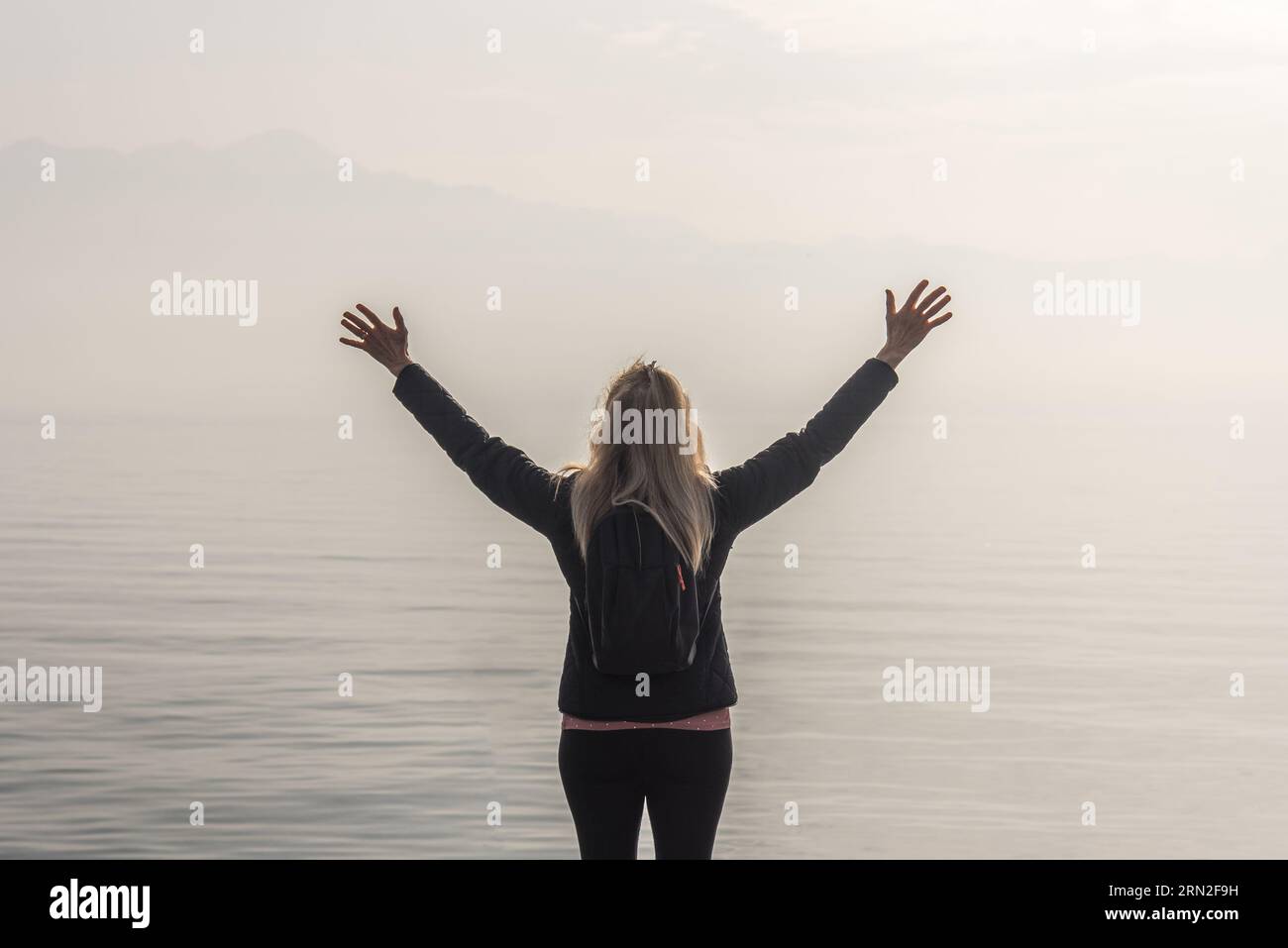 Donna viaggiatrice con le braccia alzate in piedi di fronte a un lago in una giornata nebbiosa. Foto Stock