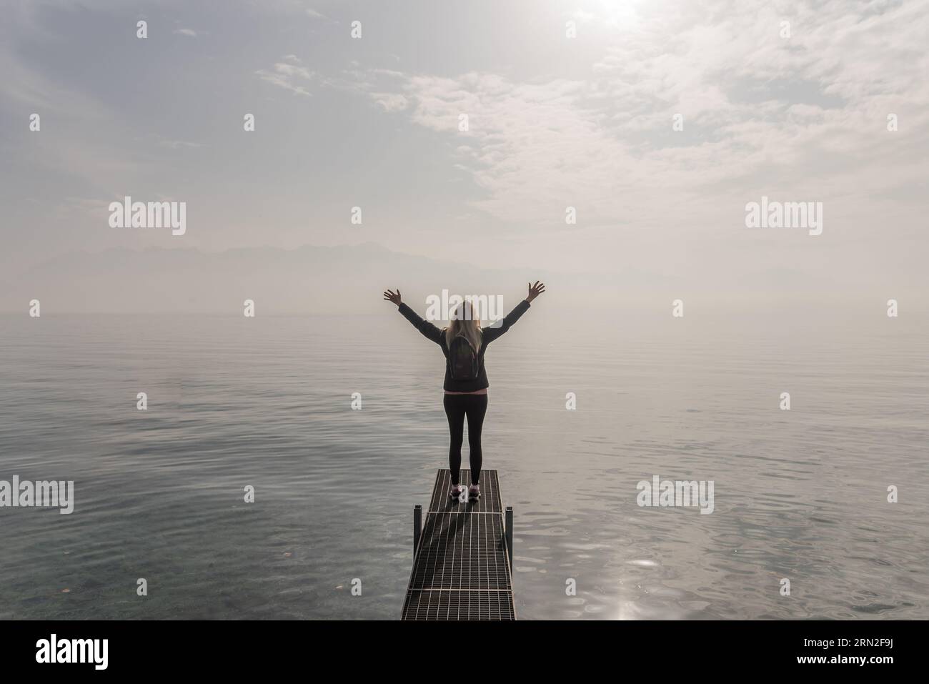 Donna viaggiatrice con le braccia alzate mentre si trova sul molo di un lago in una giornata nebbiosa. Foto Stock