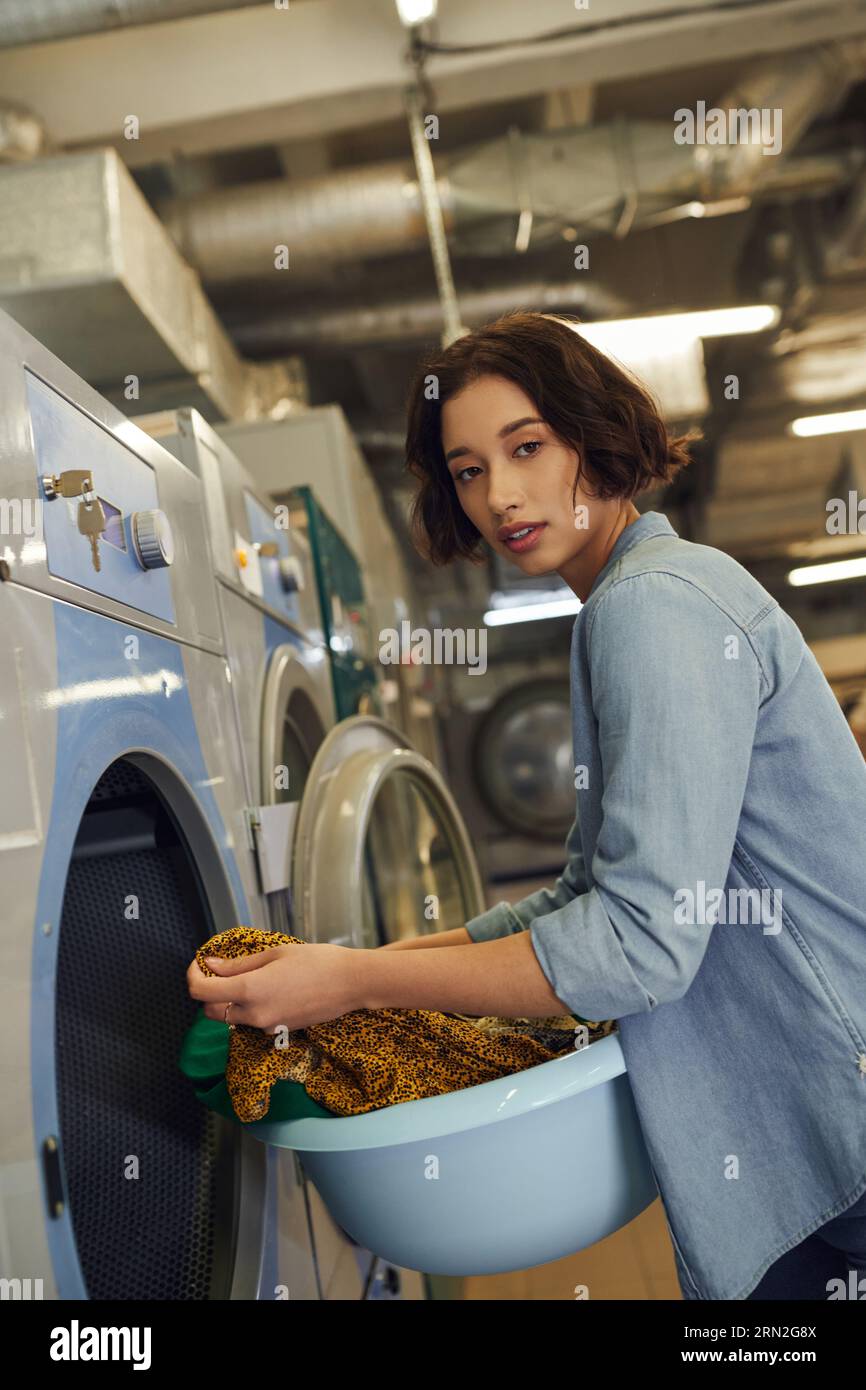 giovane donna asiatica che guarda la macchina fotografica e tiene il cesto con i vestiti nella lavanderia pubblica Foto Stock