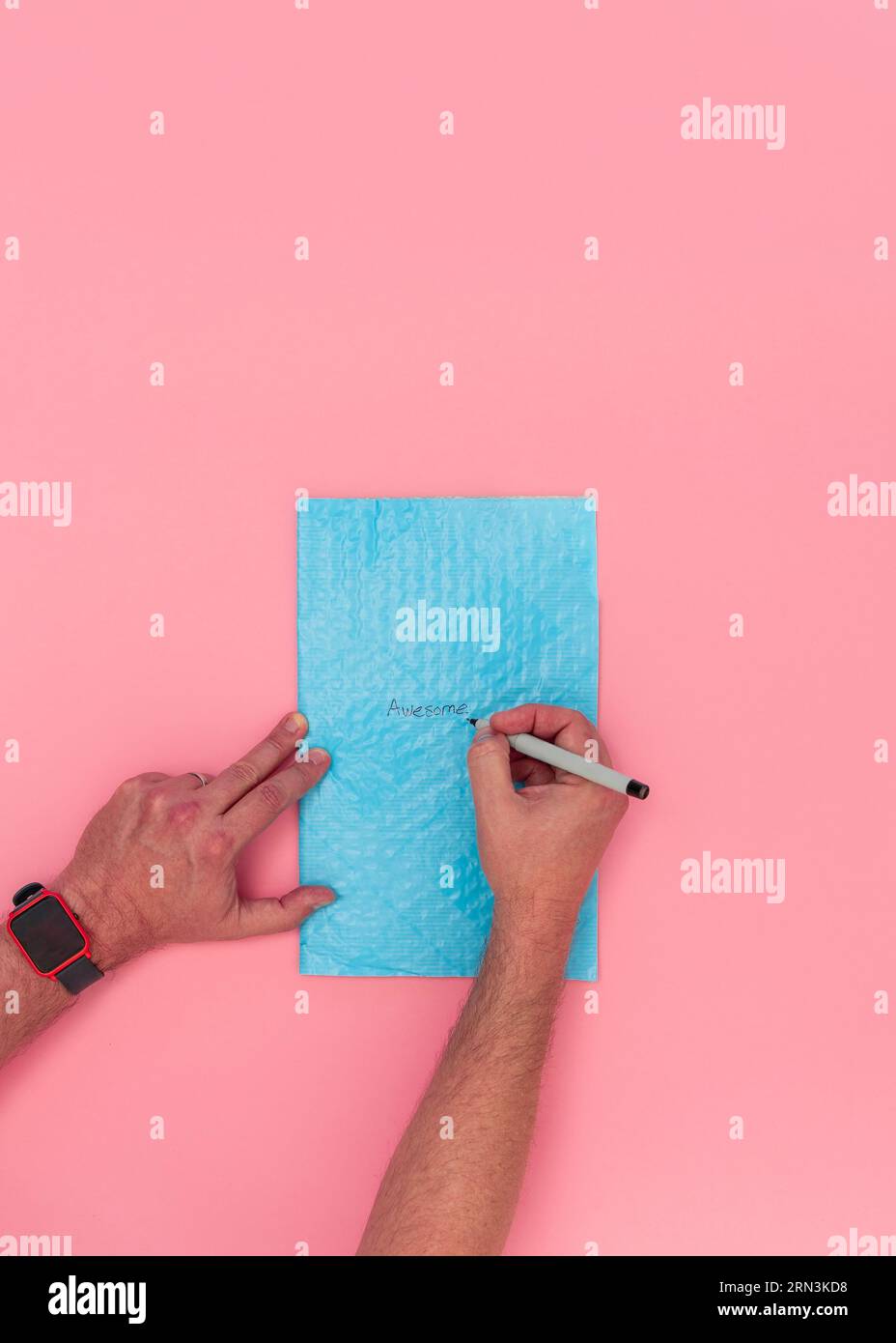 Vista dall'alto delle mani maschili adulti caucasiche che scrivono la parola "impressionante" su una busta blu a bolle d'aria. Sfondo rosa con spazio per la copia. Foto Stock