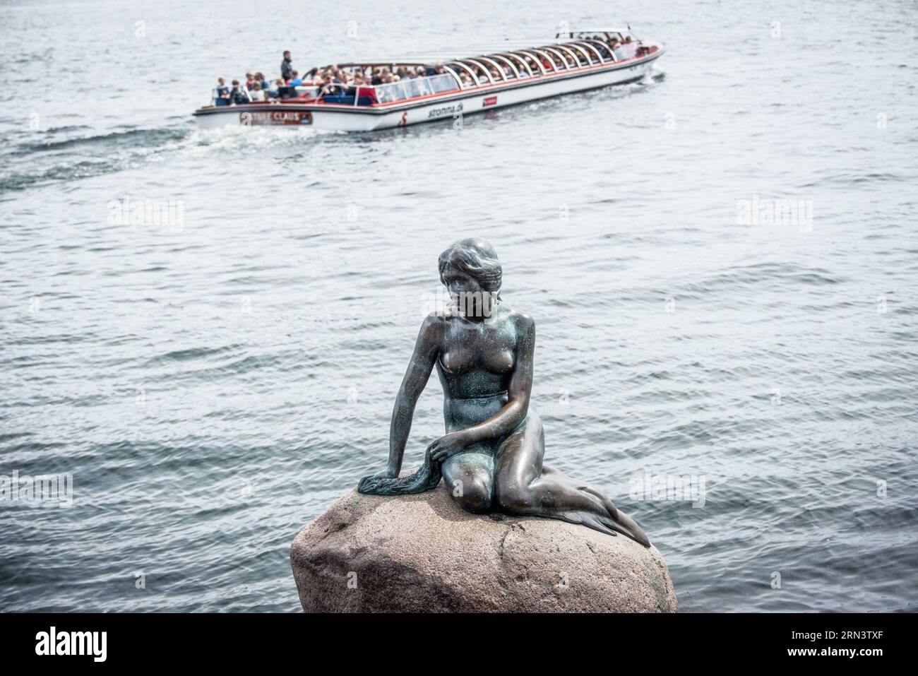 La statua della Sirenetta in bronzo Copenaghen Danimarca | COPENHAGEN, Danimarca — La statua della Sirenetta, una figura in bronzo alta circa 1,25 metri (4,1 piedi), si trova su un masso di granito alla passeggiata di Langelinie. Creata dallo scultore Edvard Eriksen e commissionata nel 1909 dal birraio Carl Jacobsen, l'opera è stata installata il 23 agosto 1913 ed è ispirata alla fiaba omonima del 1837 di Hans Christian Andersen. Qui è vista da un battello turistico di passaggio, lo "Store Claus" gestito da Stromma, offrendo una prospettiva che riflette l'intento originale della statua — essere riletta Foto Stock