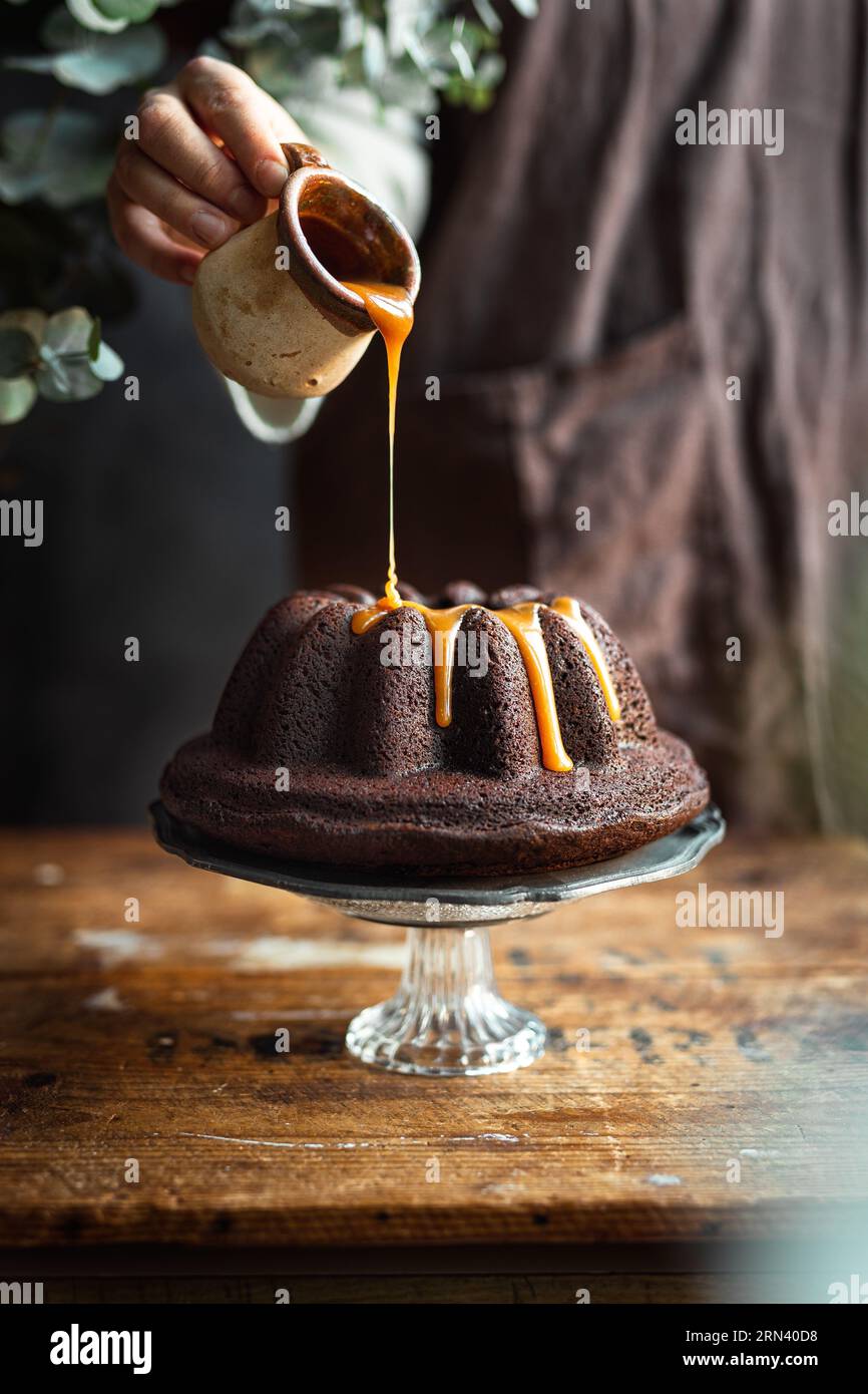 Persona sullo sfondo che versa caramello sulla torta al cioccolato. Sistemazione rustica. La torta si trova sull'elegante banco di vetro su un vecchio tavolo di legno. Foto Stock Persona sullo sfondo che versa caramello sulla torta al cioccolato. Sistemazione rustica. La torta si trova sull'elegante banco di vetro su un vecchio tavolo di legno. Foto Stock