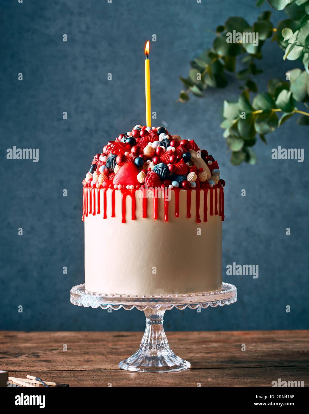 Torta di compleanno cremosa con gocce rosse e molte caramelle e frutti di bosco. In piedi su un elegante banco per dolci in vetro su un tavolo rustico in legno. Foto Stock
