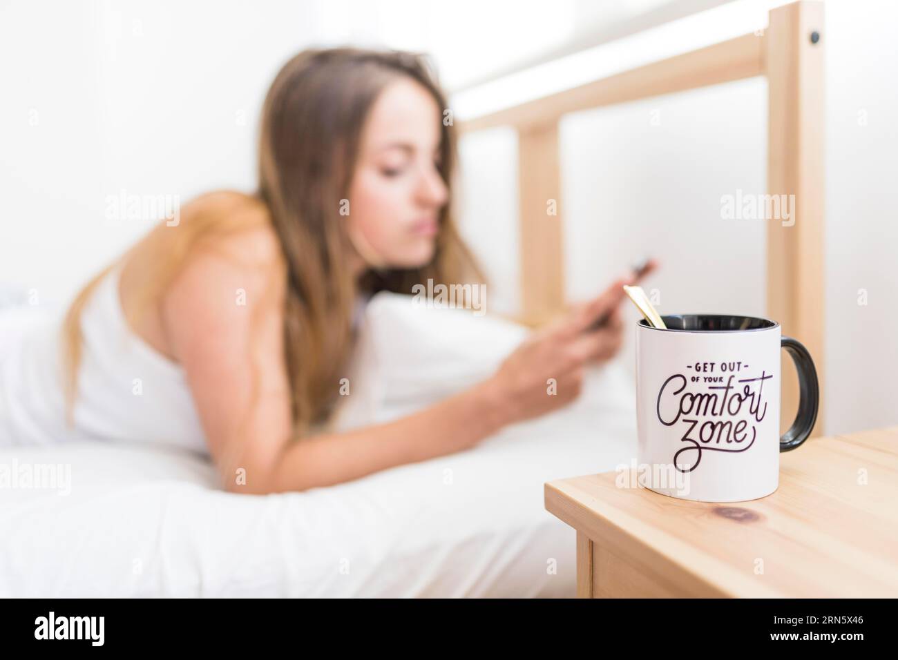 Tazza di caffè, scrivania in legno, donna sdraiata sul letto usando il telefono cellulare Foto Stock