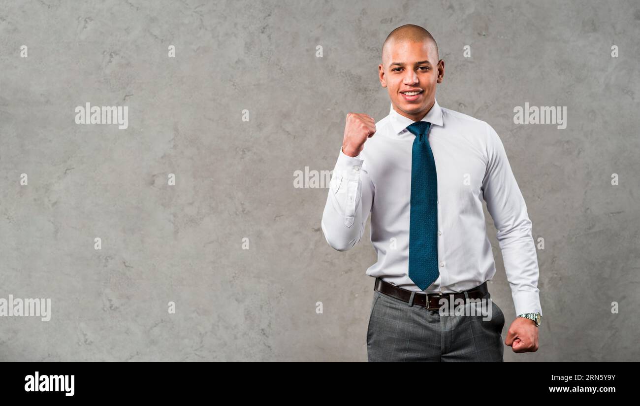 Ritratto giovane uomo d'affari sorridente in piedi contro una parete grigia che stringe il pugno Foto Stock