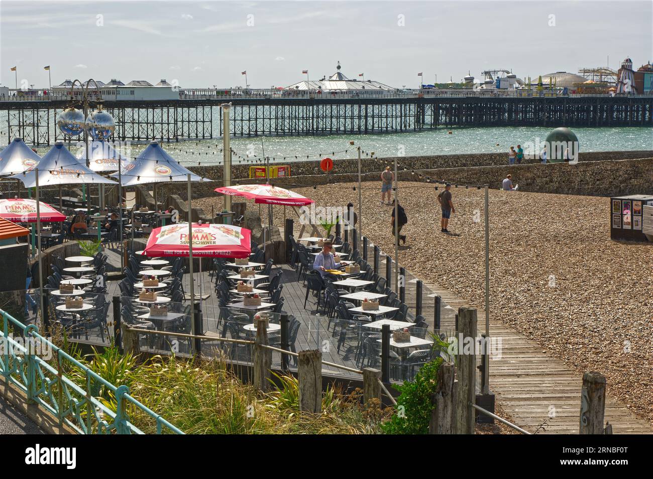 Terrace Cafe, bar e ristorante sulla spiaggia di Brighton nell'East Sussex, Inghilterra. Con Palace Pier sullo sfondo e gente che si diverte al mare. Foto Stock