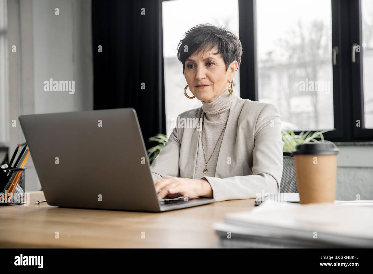 donna matura imprenditrice che digita un notebook vicino alla bevanda da asporto in un ambiente di lavoro moderno Foto Stock