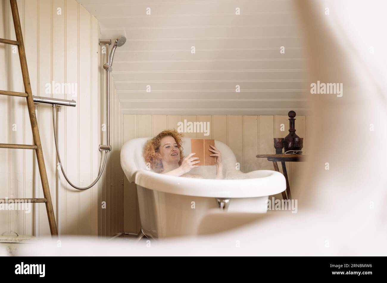 Giovane donna sdraiata nella vasca da bagno e leggendo un libro alla luce del giorno Foto Stock
