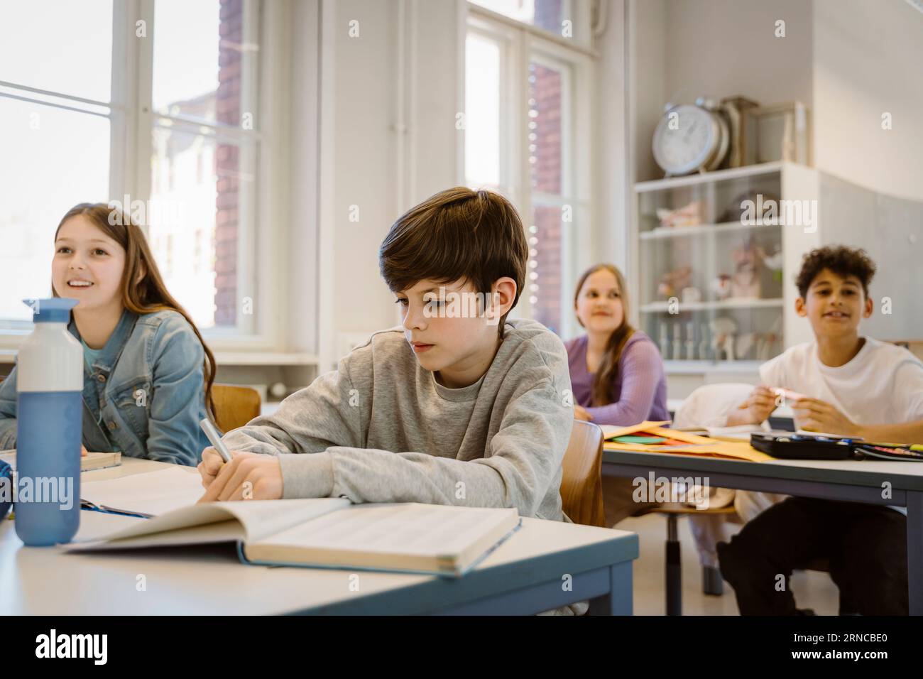 Scolaro che scrive un libro mentre è seduto alla scrivania con gli amici in classe Foto Stock