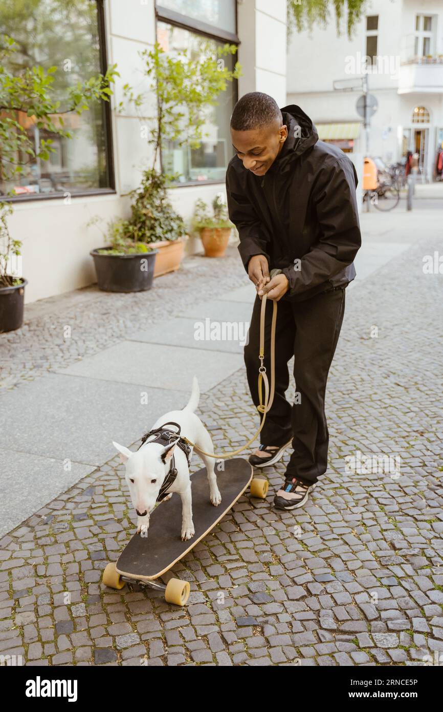 Un giovane sorridente che cammina con Bull Terrier sullo skateboard sul marciapiede Foto Stock