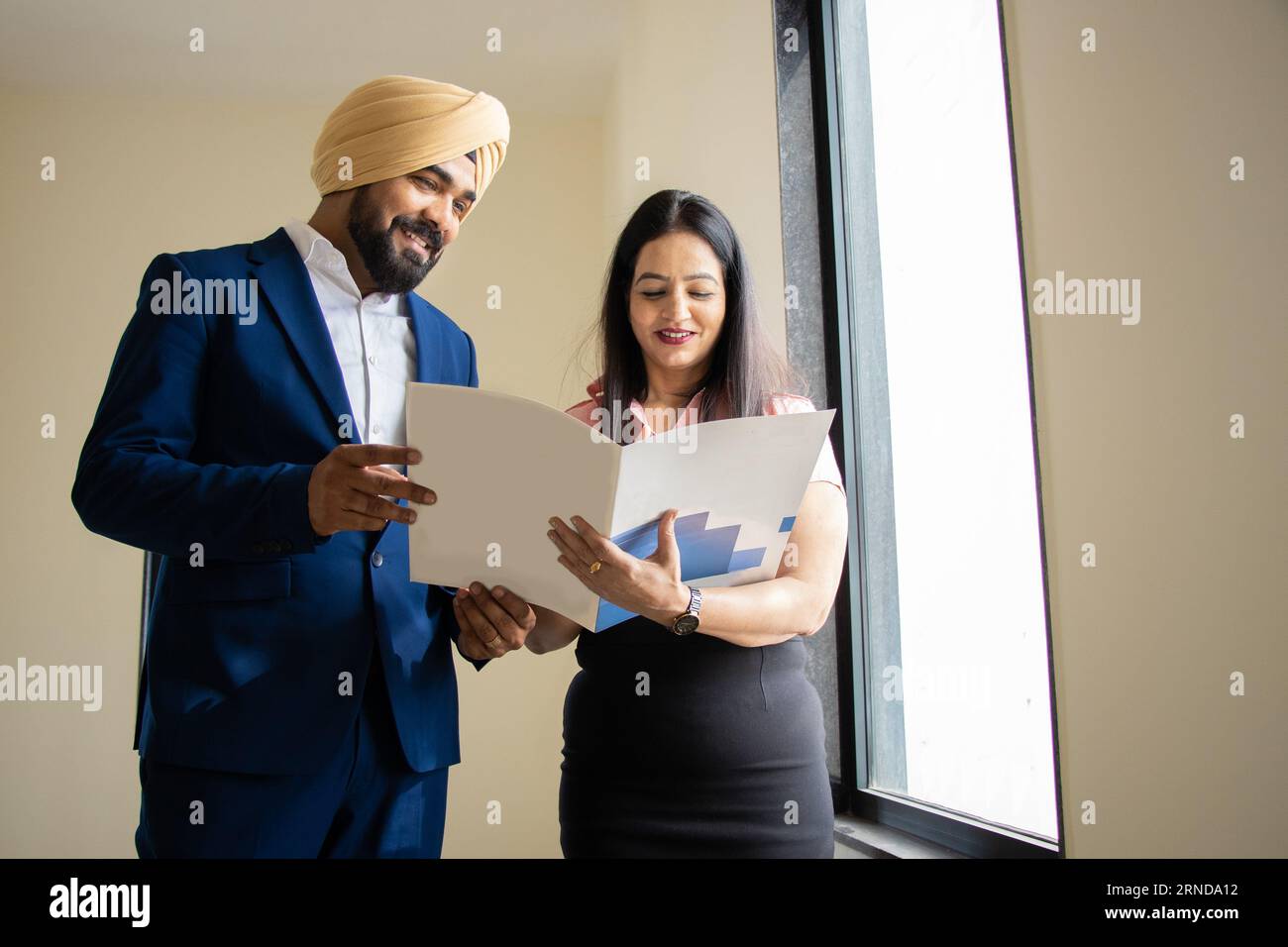 Un uomo d'affari indiano sikh che indossa un abito formale discute di lavoro con una giovane segretaria che tiene un file nell'ufficio aziendale. Foto Stock