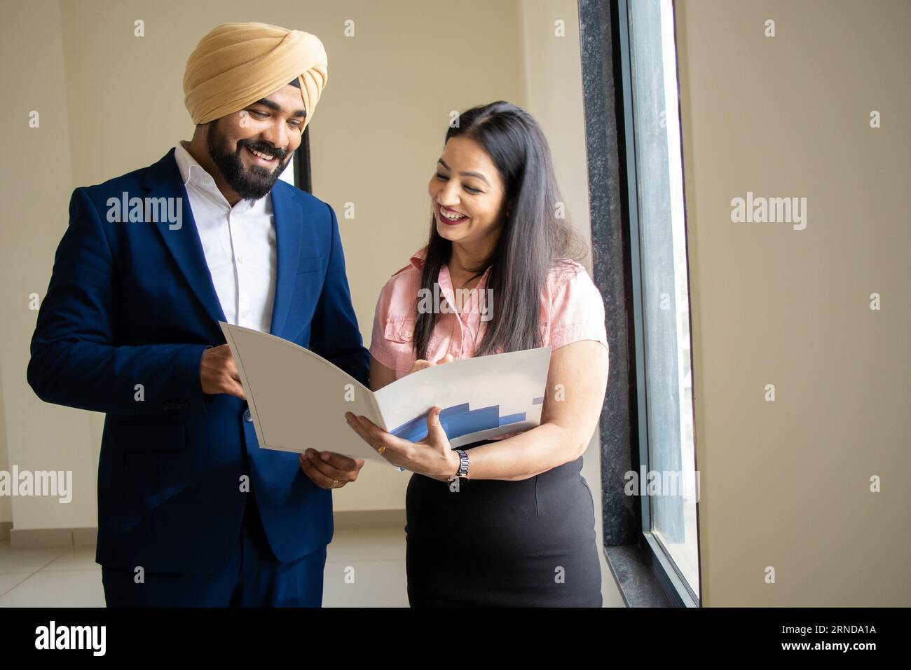 Un uomo d'affari indiano sikh che indossa un abito formale discute di lavoro con una giovane segretaria che tiene un file nell'ufficio aziendale. Foto Stock