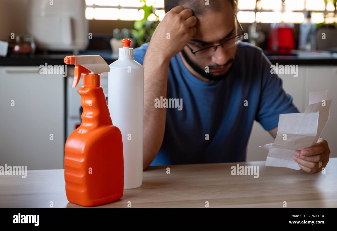 l'uomo seduto in cucina con ricevuta si sente frustrato a causa dell'inflazione e dell'aumento delle necessità quotidiane di rifornimenti per la casa con i detergenti davanti Foto Stock