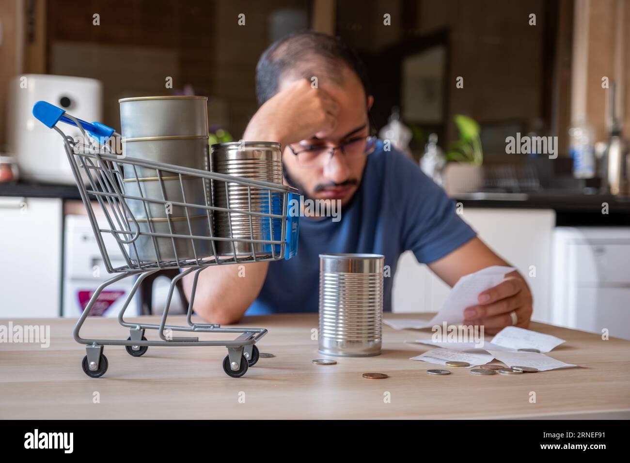un uomo seduto in cucina che tiene in mano la ricevuta sentendosi frustrato a causa dell'inflazione e dell'aumento dei bisogni quotidiani delle provviste per la casa con il carrello davanti Foto Stock