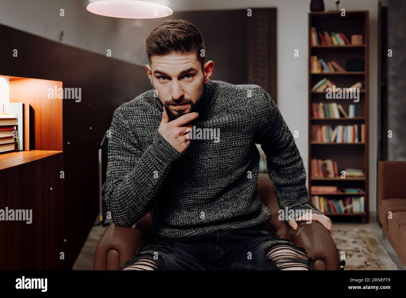 il giovane si siede in biblioteca a casa con i libri negli scaffali sullo sfondo mentre guarda la fotocamera con le dita sul mento e l'espressione scettica del volto Foto Stock