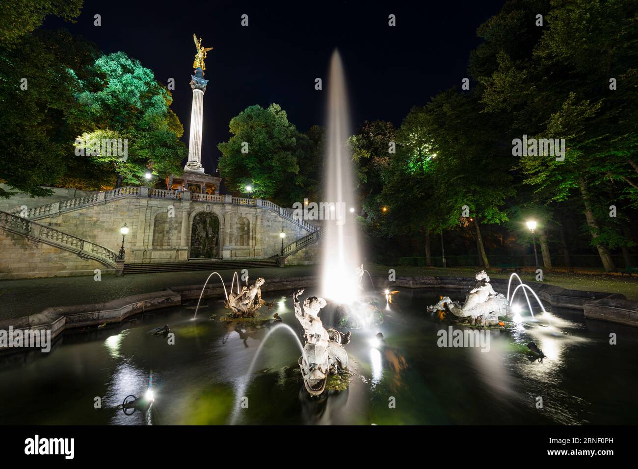 Fontana con getti d'acqua, sculture, scale e pilastro con l'angelo della pace nell'illuminazione notturna, Bogenhausen, Monaco, Germania Foto Stock