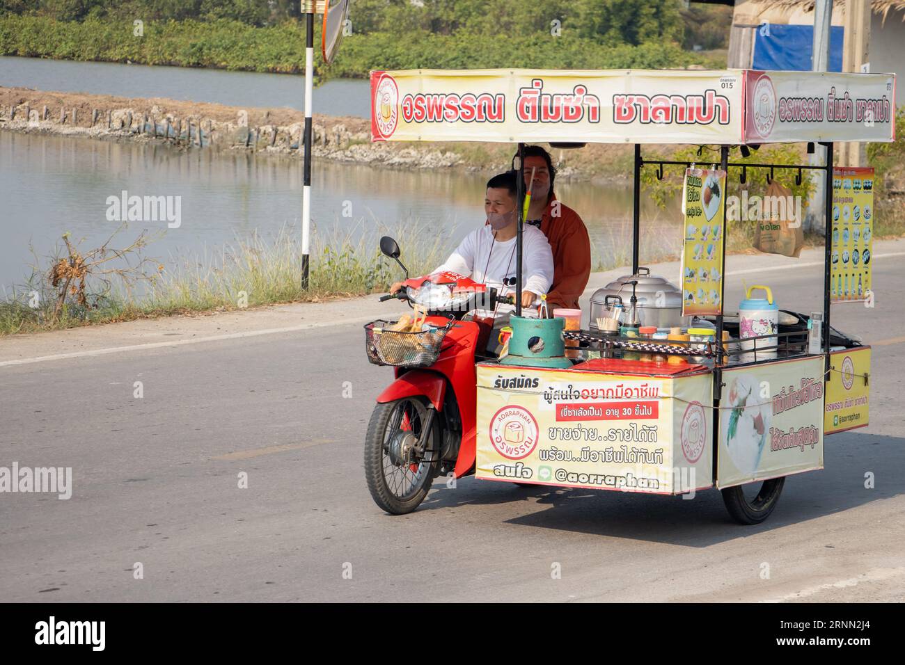 SAMUT PRAKAN, THAILANDIA, 01 2023 FEBBRAIO, Un venditore di cibo tailandese con gnocchi cavalca una motocicletta a tre ruote lungo una strada del villaggio, Thailandia Foto Stock